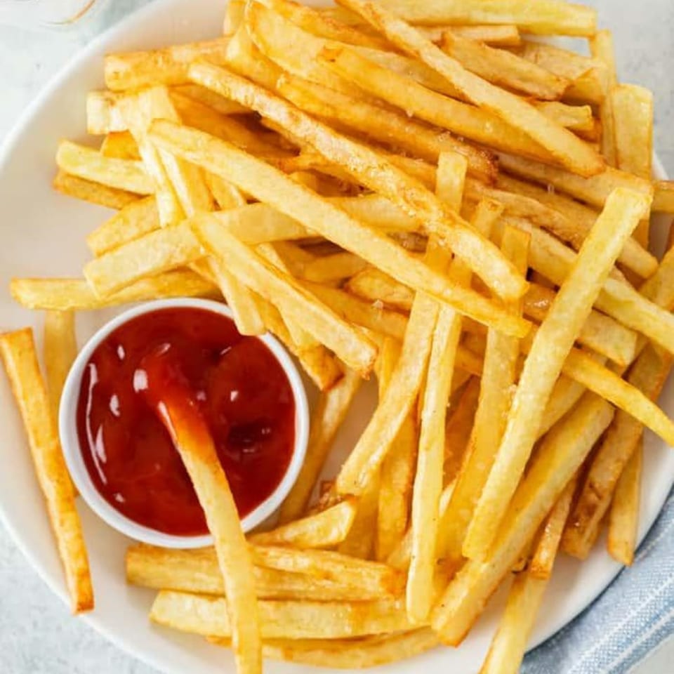 Golden french fries piled on a white plate with a side of ketchup, one fry dipped in sauce.