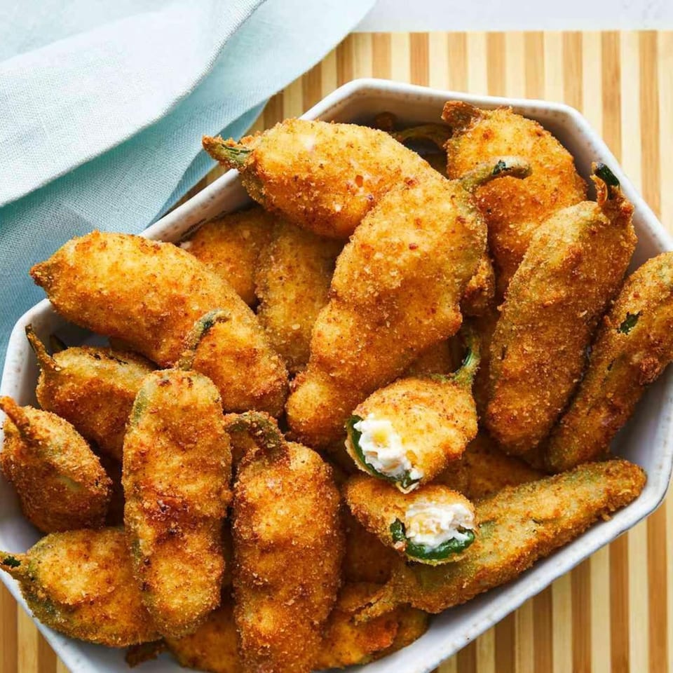 A white bowl filled with golden-brown, breaded jalapeño poppers, one cut open to show cream cheese.