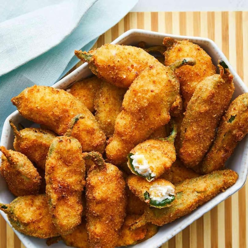 A white bowl filled with golden-brown, breaded jalapeño poppers, one cut open to show cream cheese.