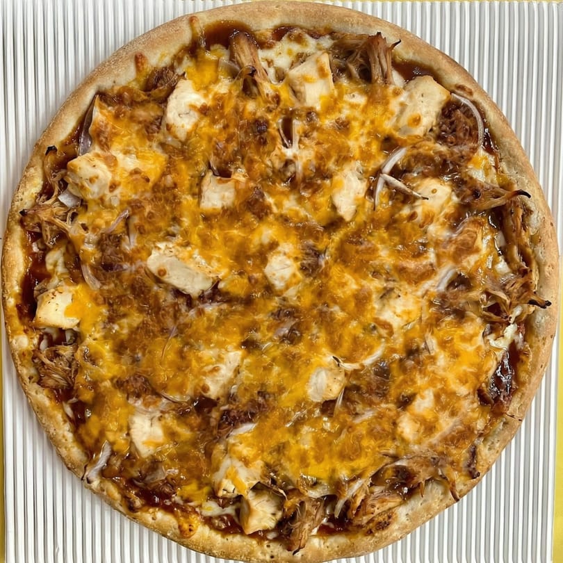 A whole BBQ chicken pizza with melted cheddar cheese, chicken pieces, and onions on a white ribbed surface.